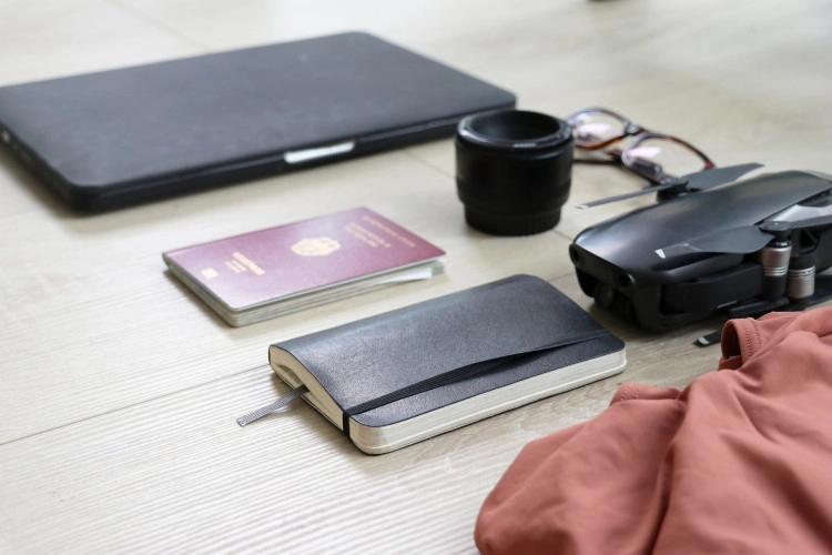 Equipo de viaje sobre una mesa: portátil, pasaporte, cuaderno, gafas y dron compacto.