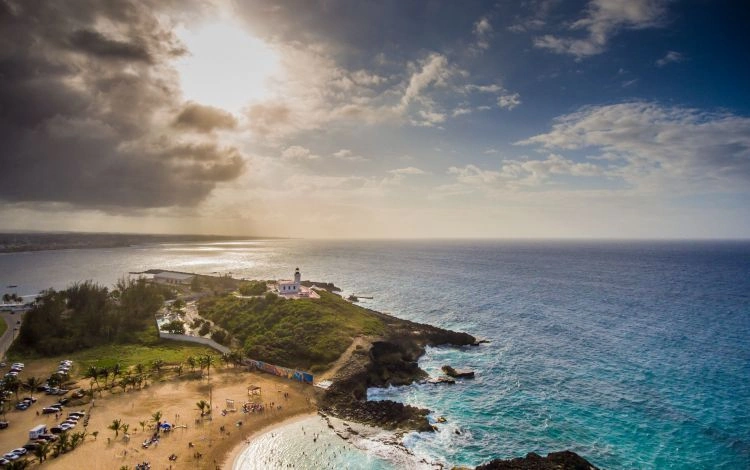 Vue aérienne d'un littoral avec une plage, un phare sur une colline et la mer sous un ciel partiellement nuageux.